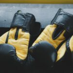 Dois pares de luvas de boxe pretas e amarelas posicionadas sobre um banco de madeira em processo de secagem natural após limpeza