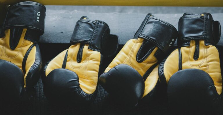 Dois pares de luvas de boxe pretas e amarelas posicionadas sobre um banco de madeira em processo de secagem natural após limpeza